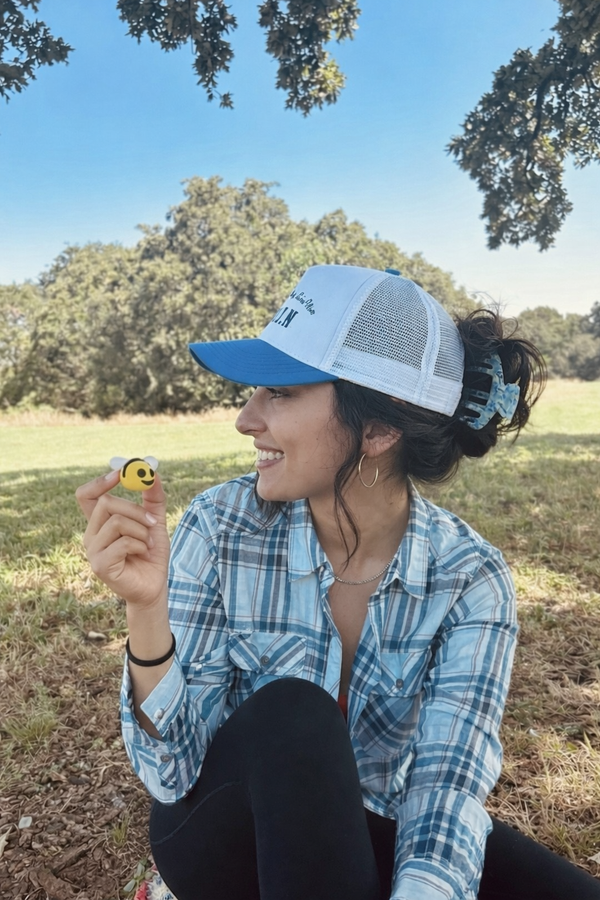 Woman sitting outdoors in a blue and white cap, hair secured in a bun with an Essential Hold claw clip in Sea Glass, wearing a plaid shirt