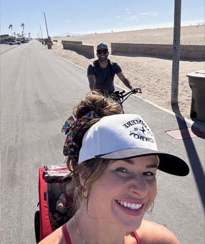 Woman wearing a white "Cowboy" graphic cap, hair pulled into a bun with an Essential Hold claw clip in Mosaic, biking on a beachside path