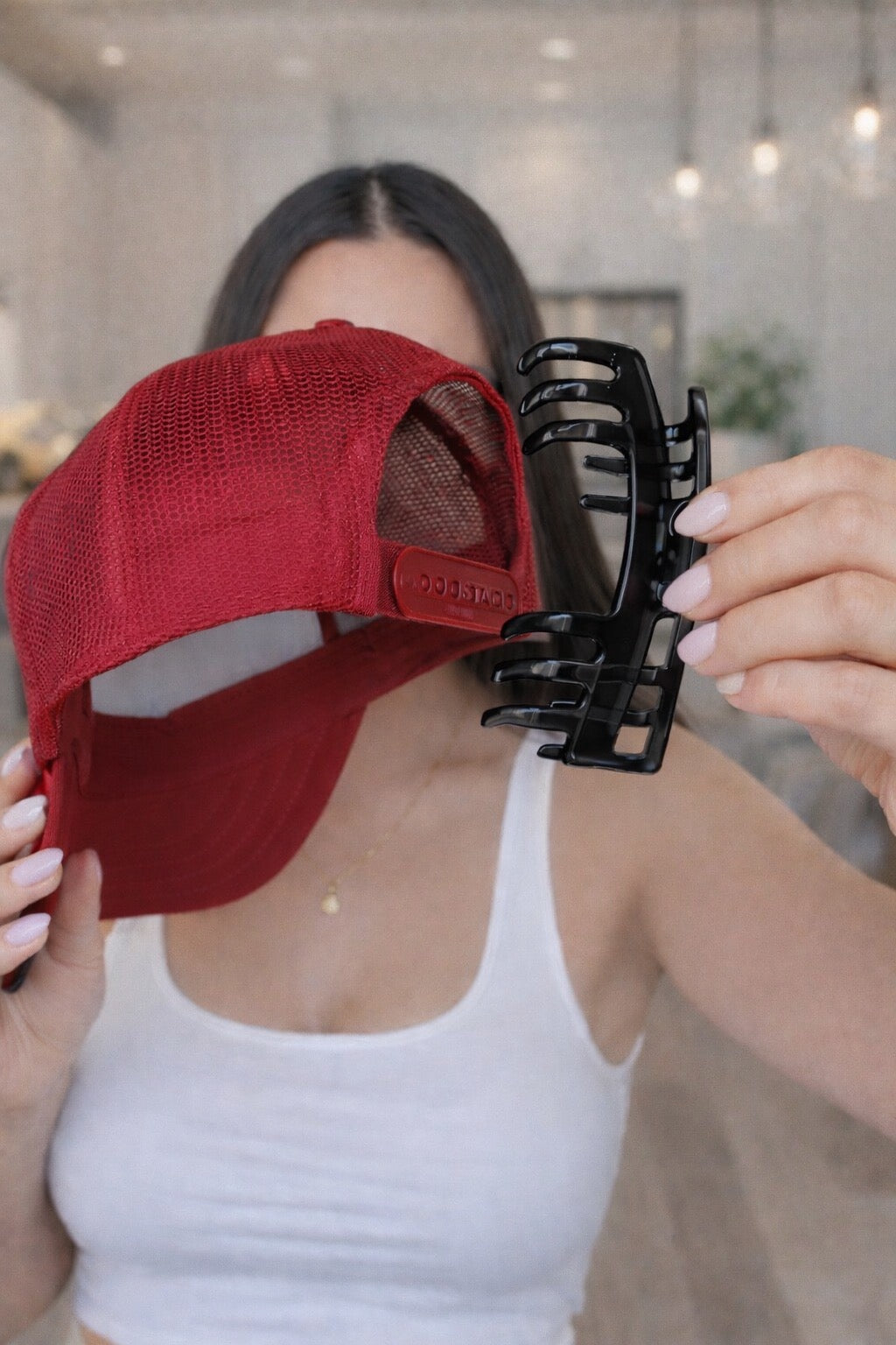 Woman holding a black Baddie Clip hat claw clip beside a red trucker hat, showing how the clip fits with the hat's adjustable back strap.

