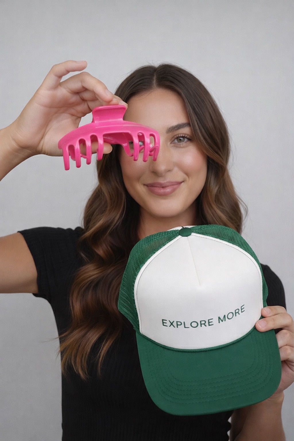 Woman holding a hot pink Baddie Clip hat claw clip beside a green and white Explore More trucker hat, showing the clip's low-profile shape.

